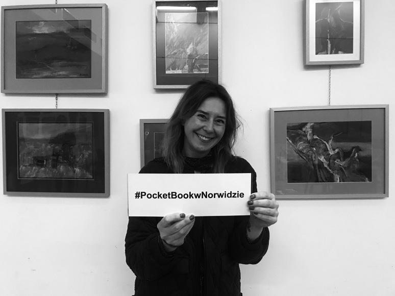 Anna Król pozuje do zdjęcia na tle obrazów. W rękach trzyma tabliczkę z napisem #PocketBook.