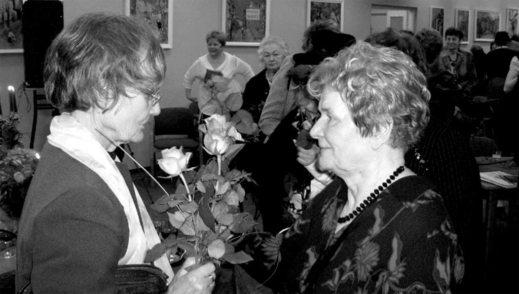 Dwie starsze kobiety stoją na przeciwko siebie. Jedna wręcza kwiaty drugiej. W tle stoją ludzie.