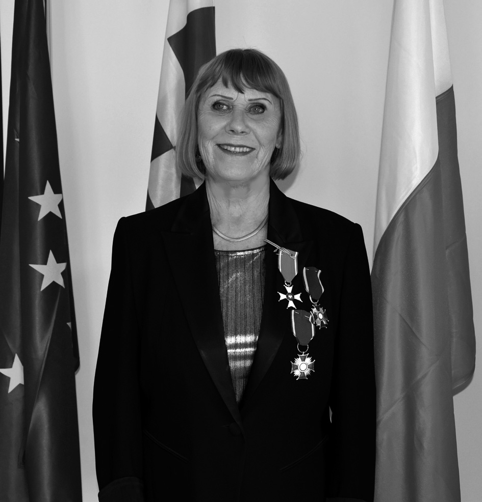 Helena Zadrejko stoi elegancko ubrana na tle trzech flag, w tym Polski i Unii Europejskiej. Na prawej piersi ma przypięte trzy medale.