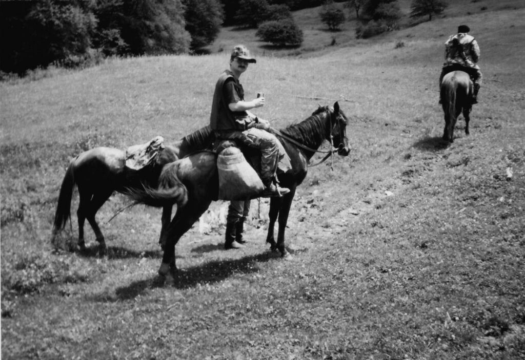 Na zdjęciu znajdują się dwie postacie jadące na koniach trzy konie. Po bokach mają bagaże. Są w lesie.